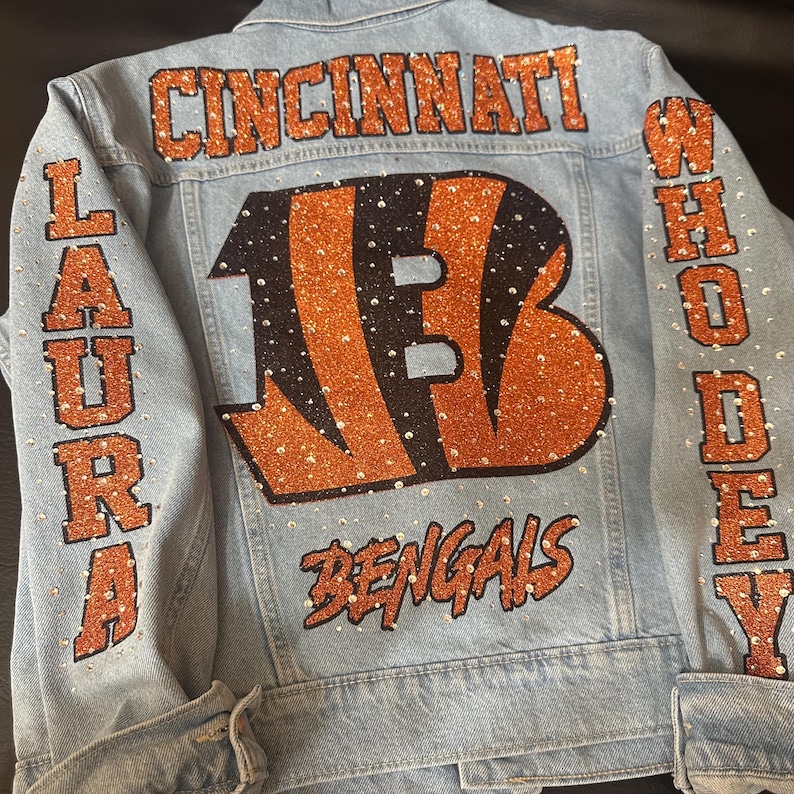 May include: A light blue denim jacket featuring orange glitter lettering that reads CINCINNATI, LAURA, WHO DEY, and BENGALS. A large orange glitter B logo is centred on the back, and the entire design is embellished with clear rhinestones.