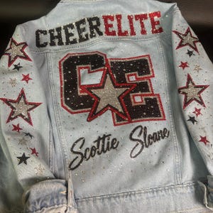 May include: A light blue denim jacket featuring the text CHEER ELITE in black and red glitter, a large CE monogram with a silver star, and the name Scottie Sloane in black script. The sleeves and back are decorated with silver, red, and black stars.