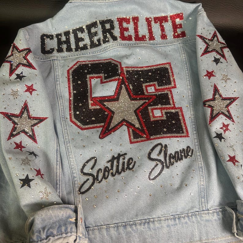 May include: A light blue denim jacket featuring the text CHEER ELITE in black and red glitter, a large CE monogram with a silver star, and the name Scottie Sloane in black script. The sleeves and back are decorated with silver, red, and black stars.