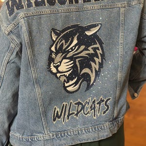May include: A light blue denim jacket featuring the text WILSONVILLE in dark blue glitter at the top, a large graphic of a wildcat head in dark blue and silver glitter in the centre, and the word WILDCATS in dark blue glitter at the bottom.