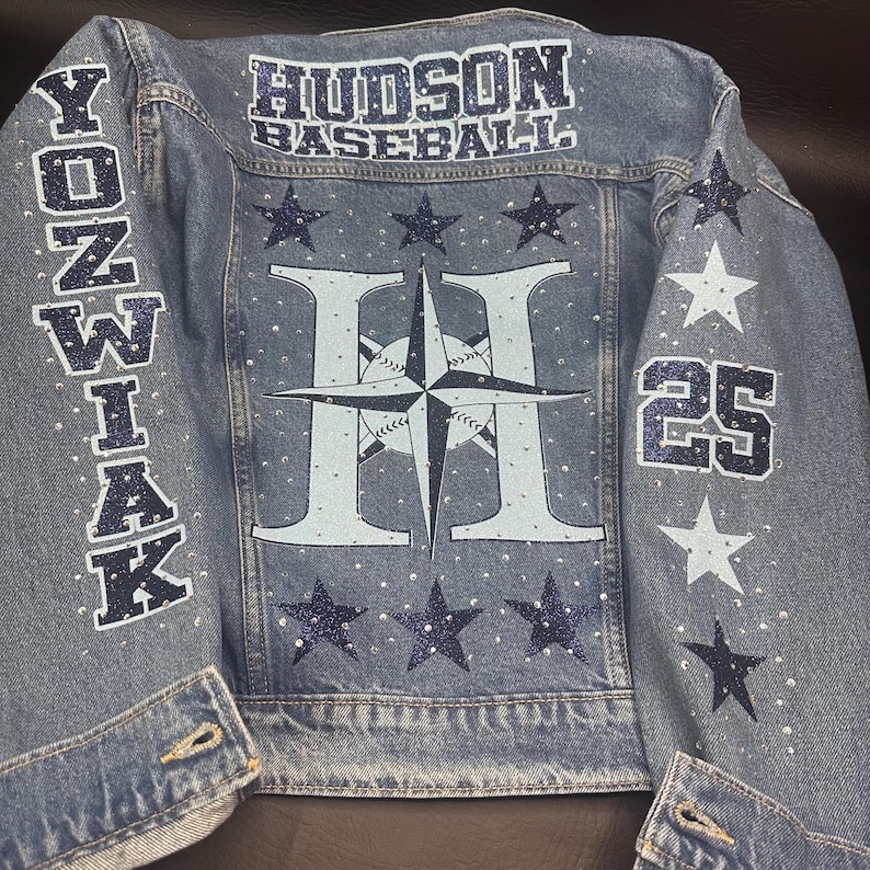 May include: A light blue denim jacket featuring the text HUDSON BASEBALL at the top, YONZWIAK on the left sleeve, and the number 25 on the right sleeve. The back displays a large letter H with a compass star and baseball graphic.