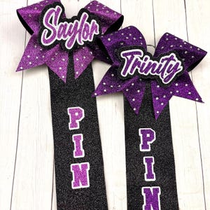 Puede incluir: Dos lazos de animadora de purpurina negra y morada con las palabras "Saylor" y "Trinity" en ellos. Los lazos están unidos a cintas de purpurina negra y morada con la palabra "PIN" escrita verticalmente en ellos.