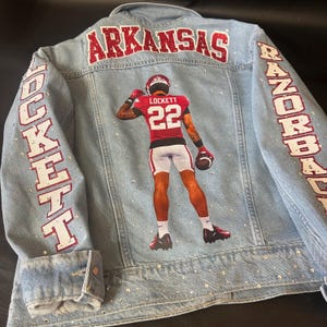 May include: A light blue denim jacket featuring a graphic of a football player wearing a red jersey with the name LOCKETT and number 22. The word ARKANSAS is in red glitter across the top, with LOCKETT and RAZORBACKS on the sleeves.