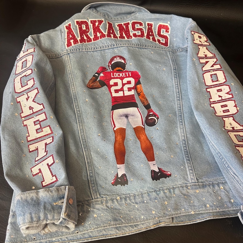 May include: A light blue denim jacket featuring a graphic of a football player wearing a red jersey with the name LOCKETT and number 22. The word ARKANSAS is in red glitter across the top, with LOCKETT and RAZORBACKS on the sleeves.