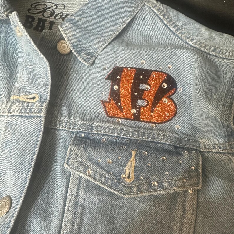 May include: A light blue denim jacket featuring a glittery orange and black letter B logo on the chest. The jacket is embellished with scattered clear rhinestones across the front pocket and around the logo. A partial label reads BENGALS.