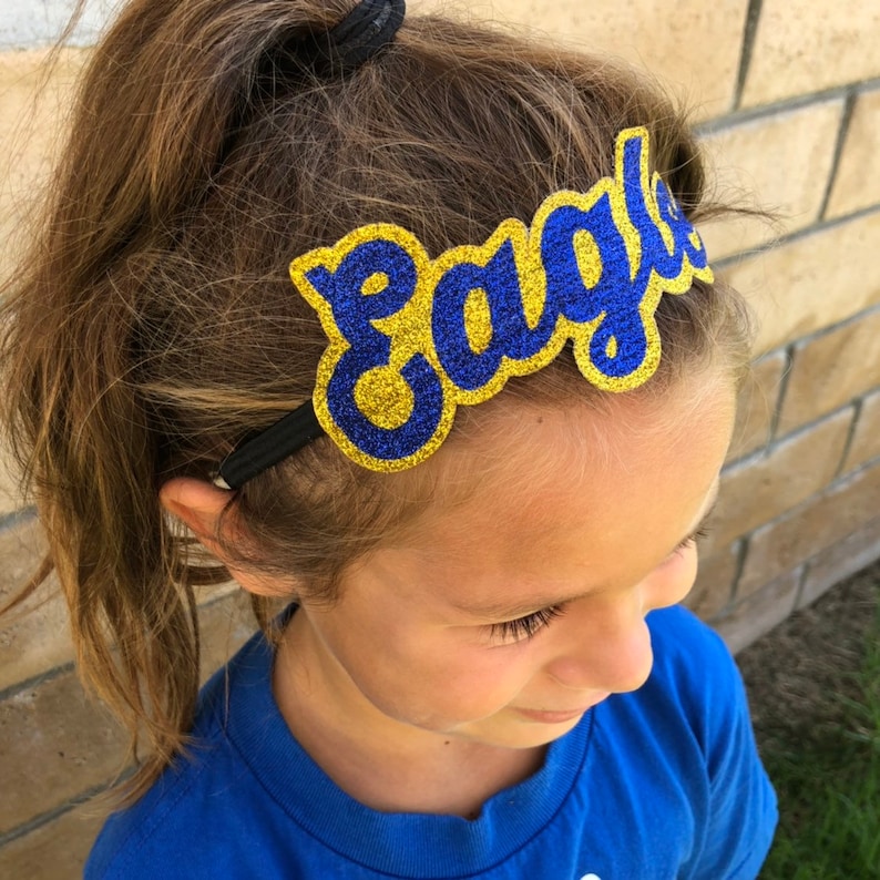 May include: A blue and gold glitter headband with the word "Eagles" on it. The headband is worn by a young person with brown hair.