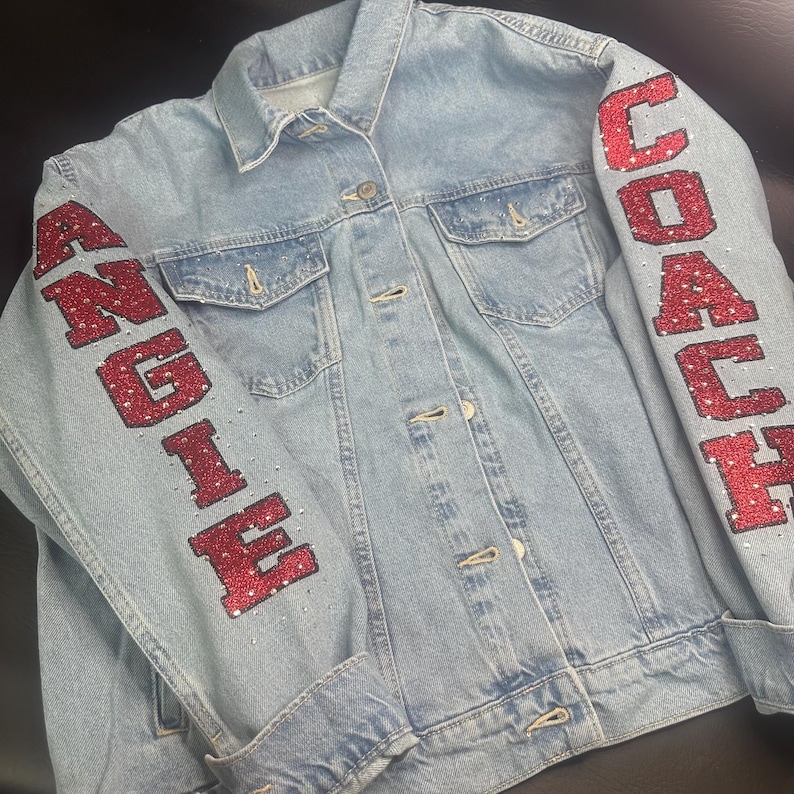 May include: A light blue denim jacket featuring red glittery lettering on the sleeves. The left sleeve displays the name ANGIE and the right sleeve displays the word COACH. Both sleeves are embellished with small clear rhinestones.