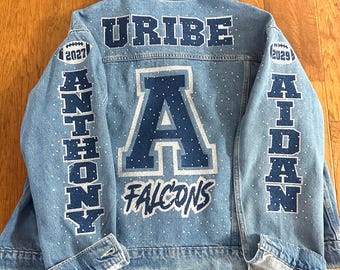 May include: A light blue denim jacket featuring dark blue and silver glitter lettering and graphics. The back displays the name URIBE, a large letter A, and the word FALCONS. The sleeves feature the names ANTHONY and AIDAN with football icons.