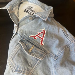 May include: A light blue denim jacket featuring a red glittery letter A patch on the chest pocket. The jacket is embellished with small clear rhinestones and has a collar. The interior label displays the text Bows and BALLETS.
