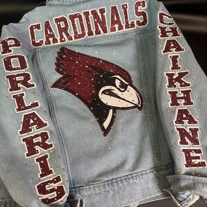 May include: A light blue denim jacket featuring a large glittery maroon cardinal bird logo on the back. The word CARDINALS is printed in maroon glitter above the logo, with PORLARIS and CHAIKHANE on the sleeves. Clear rhinestones accent the fabric.