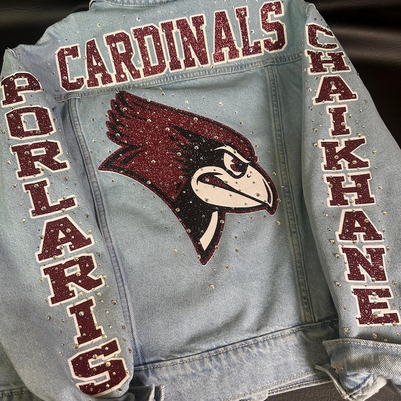 May include: A light blue denim jacket featuring a large glittery maroon cardinal bird logo on the back. The word CARDINALS is printed in maroon glitter above the logo, with PORLARIS and CHAIKHANE on the sleeves. Clear rhinestones accent the fabric.