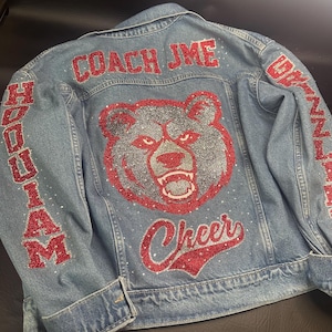 May include: A light blue denim jacket featuring red glitter text that reads COACH JME and Cheer. The back displays a large silver and red glitter bear head graphic. The sleeves feature red glitter text reading HOQUIAM and GRIZZLIES.