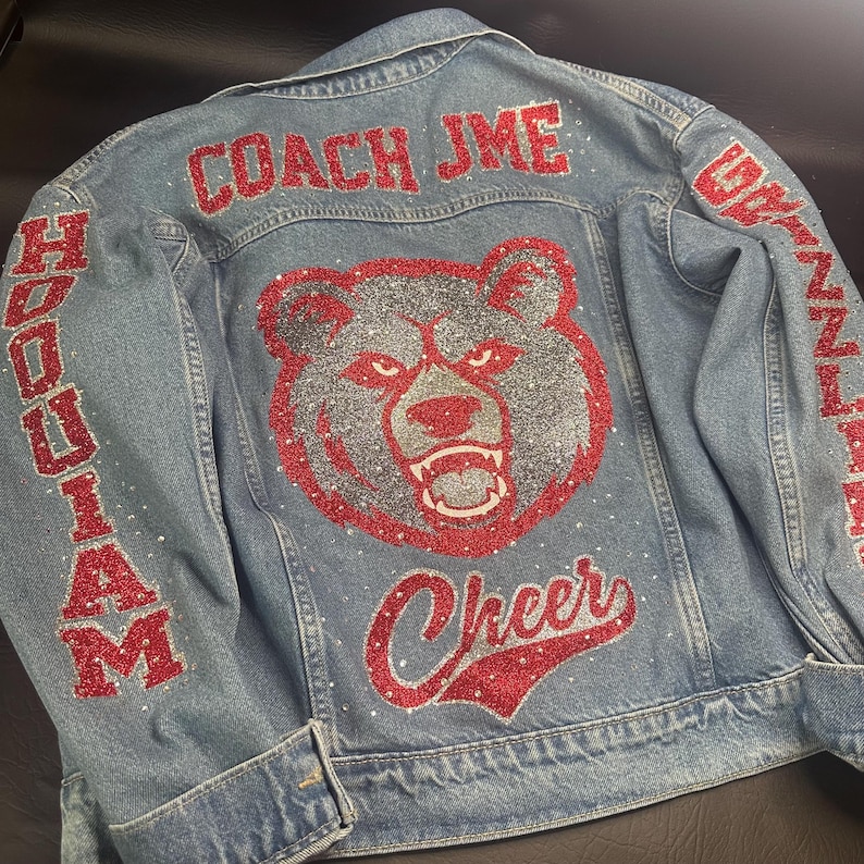 May include: A light blue denim jacket featuring red glitter text that reads COACH JME and Cheer. The back displays a large silver and red glitter bear head graphic. The sleeves feature red glitter text reading HOQUIAM and GRIZZLIES.