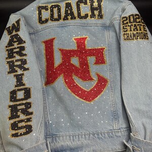 May include: A light blue denim jacket featuring the word COACH in black and gold glitter, the word WARRIORS in black and gold on the sleeve, and 2024 STATE CHAMPIONS on the other sleeve. A large red and gold glitter logo sits in the center.