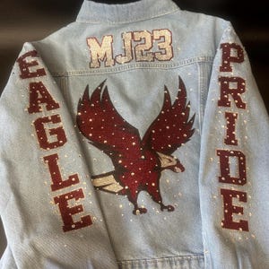 May include: A light blue denim jacket featuring a large dark red glitter eagle graphic on the back. The text MJ23 is in silver glitter above the eagle. The sleeves feature the words EAGLE and PRIDE in dark red glitter, accented with clear rhinestones.