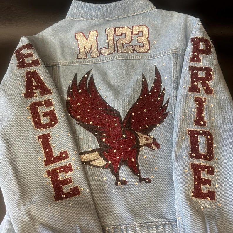 May include: A light blue denim jacket featuring a large dark red glitter eagle graphic on the back. The text MJ23 is in silver glitter above the eagle. The sleeves feature the words EAGLE and PRIDE in dark red glitter, accented with clear rhinestones.