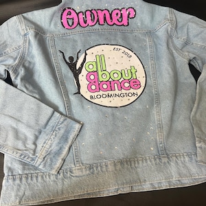 May include: A light blue denim jacket featuring the word Owner in pink letters at the top. Below, a circular graphic displays a black silhouette of a dancer, the text all about dance in green and pink, and the words EST 2018 and BLOOMINGTON.