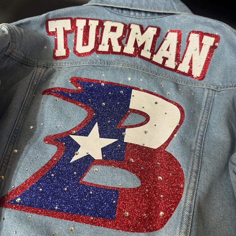 May include: A light blue denim jacket featuring the name TURMAN in white letters with red glitter outlines. Below, a large letter B is decorated with red and blue glitter, a white star, and scattered clear rhinestones across the fabric.