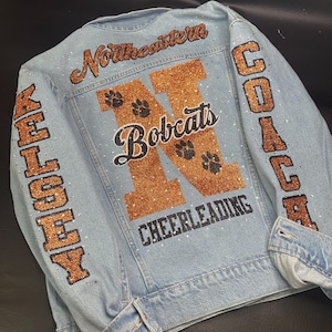 May include: A light blue denim jacket featuring orange glitter lettering that reads Northeastern, Bobcats, Cheerleading, Kelsey, and Coach. The back and sleeves are decorated with small clear rhinestones and black paw print graphics.