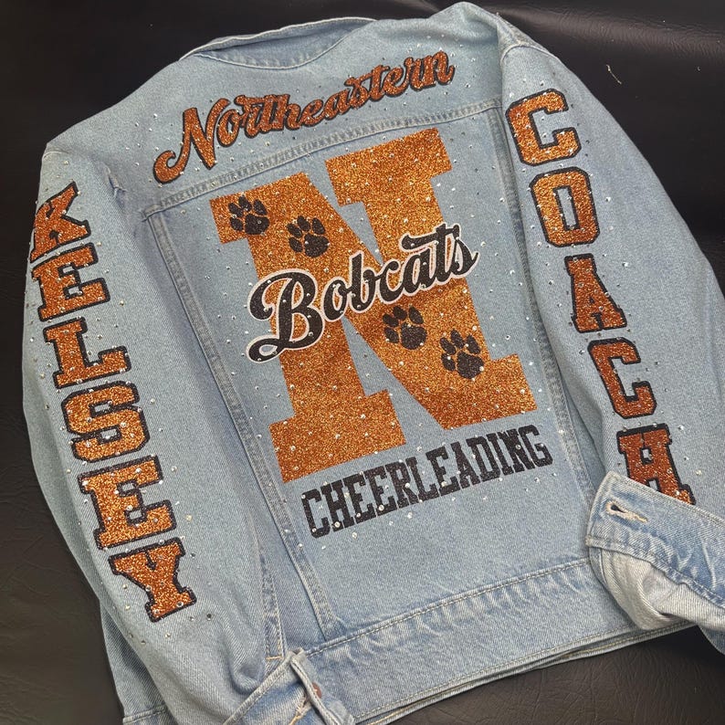 May include: A light blue denim jacket featuring orange glitter lettering that reads Northeastern, Bobcats, Cheerleading, Kelsey, and Coach. The back and sleeves are decorated with small clear rhinestones and black paw print graphics.