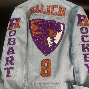 May include: A light blue denim jacket featuring purple and orange glitter lettering that reads SILICH, HOBART, and HOCKEY. The back displays a purple and orange mascot graphic and the number 8, all embellished with scattered clear rhinestones.
