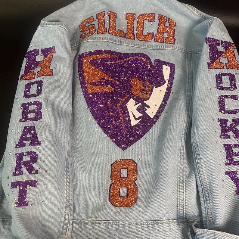 May include: A light blue denim jacket featuring purple and orange glitter lettering that reads SILICH, HOBART, and HOCKEY. The back displays a purple and orange mascot graphic and the number 8, all embellished with scattered clear rhinestones.