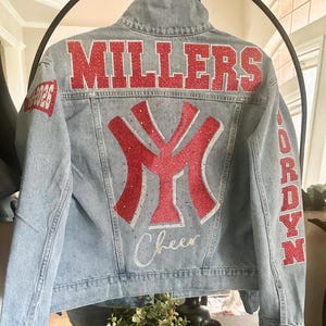 May include: A light blue denim jacket featuring red glitter lettering that reads MILLERS across the back shoulders. A large red glitter NY logo is centered on the back with the word Cheer in silver script below. JORDYN is in red on the sleeve.