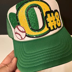 May include: Green, yellow, and black baseball cap with a white front panel. The front panel features a large green and yellow letter "O" with rhinestones, a baseball, and the number "#8" in black with rhinestones.
