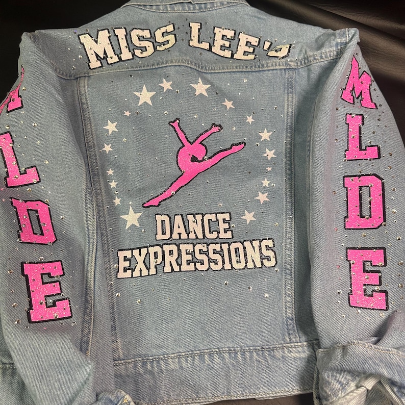 May include: A light blue denim jacket featuring the text MISS LEE'S DANCE EXPRESSIONS and a pink silhouette of a dancer. The back and sleeves are decorated with white stars and scattered rhinestones, with the letters MLDE on the sleeves.