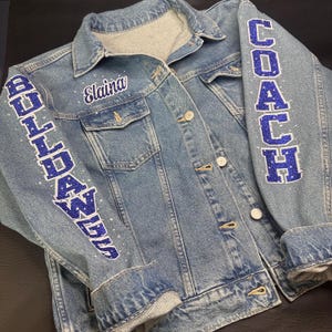 May include: A light blue denim jacket featuring the name Elaina embroidered in white on the chest. The sleeves are decorated with blue glitter lettering that reads BULLDAWGS on the left and COACH on the right, accented with small clear rhinestones.