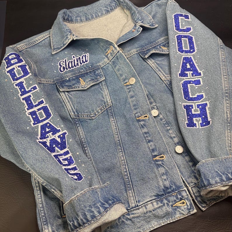 May include: A light blue denim jacket featuring the name Elaina embroidered in white on the chest. The sleeves are decorated with blue glitter lettering that reads BULLDAWGS on the left and COACH on the right, accented with small clear rhinestones.