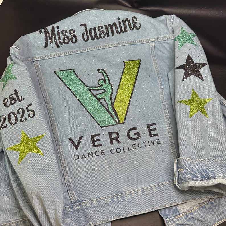May include: A light blue denim jacket featuring the text Miss Jasmine, est. 2025, and Verge Dance Collective. The back displays a large green glitter V logo with a dancer silhouette, accented by scattered rhinestones and green and black stars.