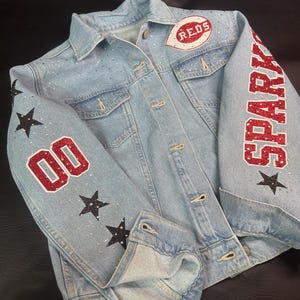May include: Light blue denim jacket with rhinestone embellishments. Features the word "SPARKLE" in red glitter on the right sleeve and the number "00" on the left sleeve. Includes a "REDS" patch on the chest and black star accents.