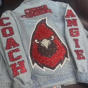 May include: A light blue denim jacket featuring red glitter lettering that reads "CHEER COMBINE" at the top, "COACH" on the left sleeve, and "ANGIE" on the right sleeve. A large red and black cardinal bird graphic is centered on the back.