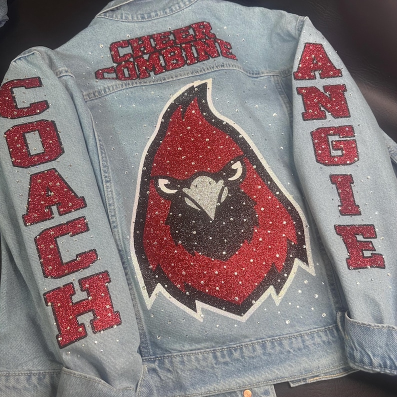 May include: A light blue denim jacket featuring red glitter lettering that reads "CHEER COMBINE" at the top, "COACH" on the left sleeve, and "ANGIE" on the right sleeve. A large red and black cardinal bird graphic is centered on the back.