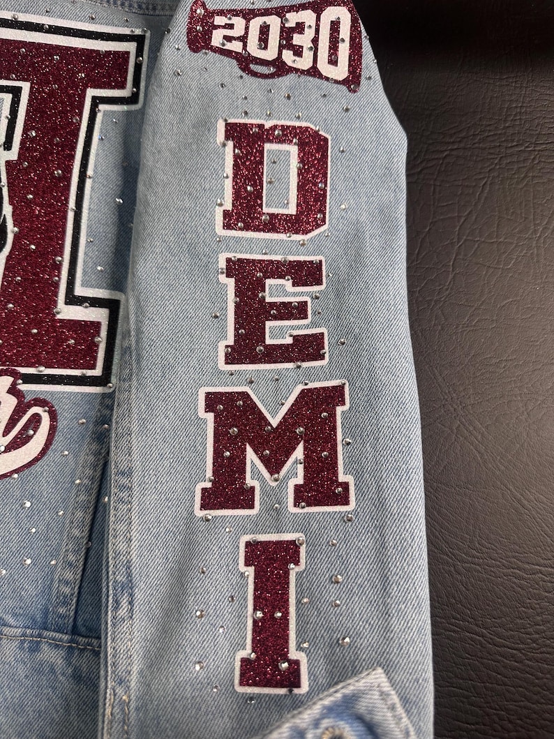 May include: A light blue denim jacket sleeve featuring the name DEMI in large, vertical, maroon glitter letters with white outlines. Above the name is a maroon glitter megaphone graphic with the text 2030. Small clear rhinestones are scattered.