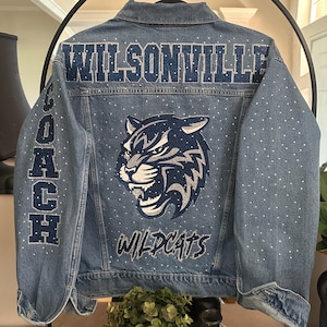 May include: A blue denim jacket featuring the text WILSONVILLE in dark blue glitter across the upper back. The word COACH is written vertically on the left sleeve. A large wildcat mascot graphic and the word WILDCATS appear below.