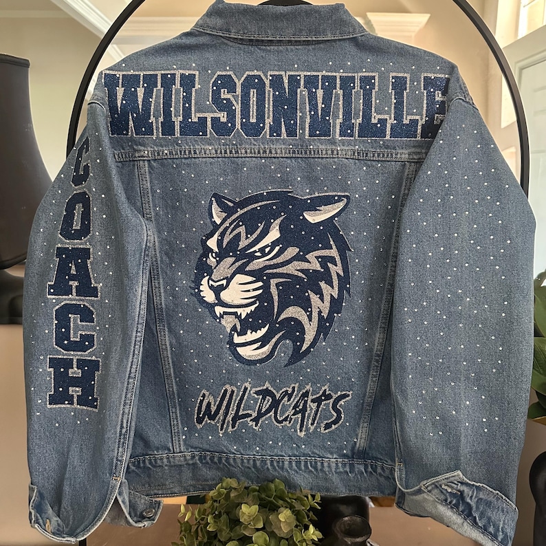 May include: A blue denim jacket featuring the text WILSONVILLE in dark blue glitter across the upper back. The word COACH is written vertically on the left sleeve. A large wildcat mascot graphic and the word WILDCATS appear below.