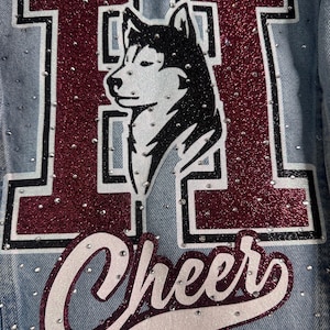 May include: A light blue denim jacket featuring a large letter H in maroon glitter with a black outline. A black and white husky dog illustration is centered within the letter. The word Cheer is written below in white glitter script with a maroon outline.