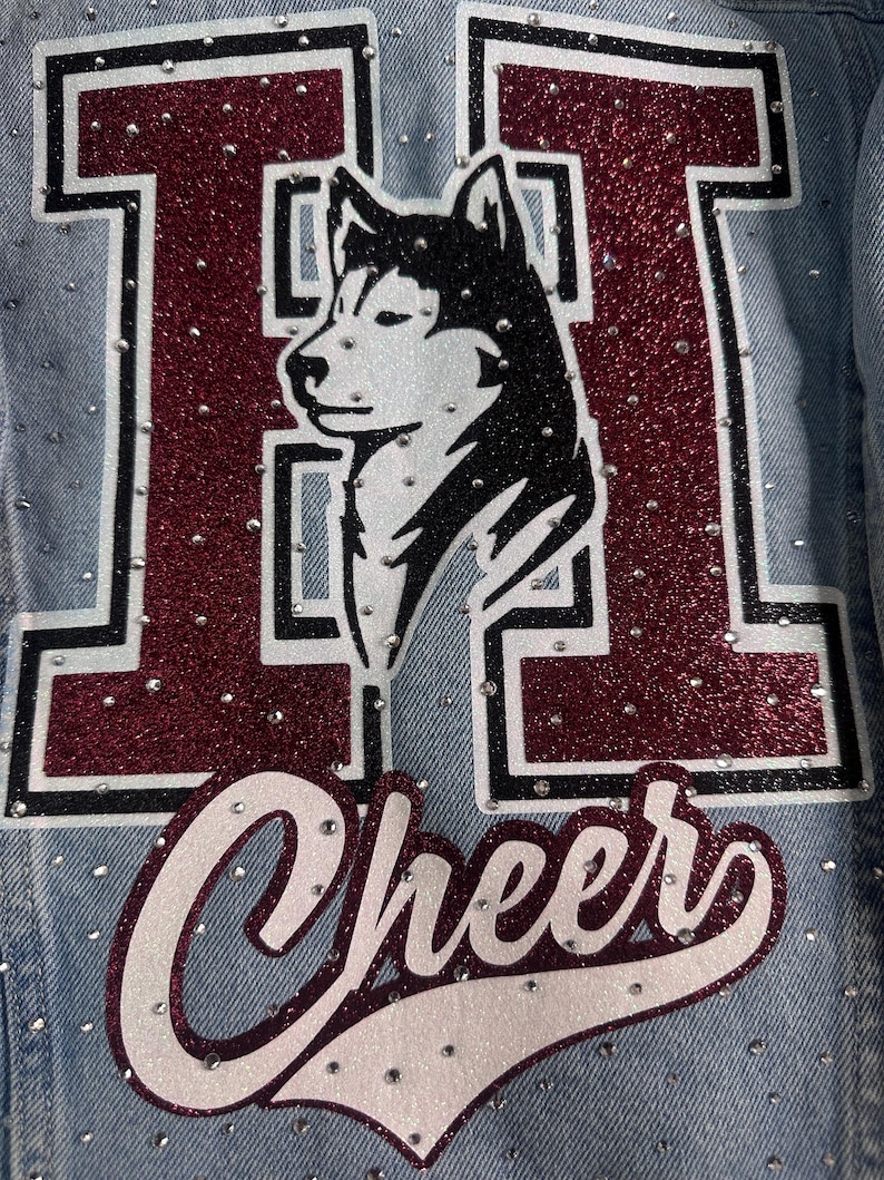 May include: A light blue denim jacket featuring a large letter H in maroon glitter with a black outline. A black and white husky dog illustration is centered within the letter. The word Cheer is written below in white glitter script with a maroon outline.