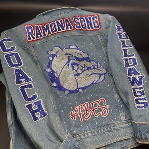 May include: A light blue denim jacket featuring a large silver and blue glitter bulldog graphic on the back. The text RAMONA SONG is in red and blue glitter, COACH and BULLDOGS are in blue glitter, and #FBES is in red glitter. Small clear rhinestones are scattered.
