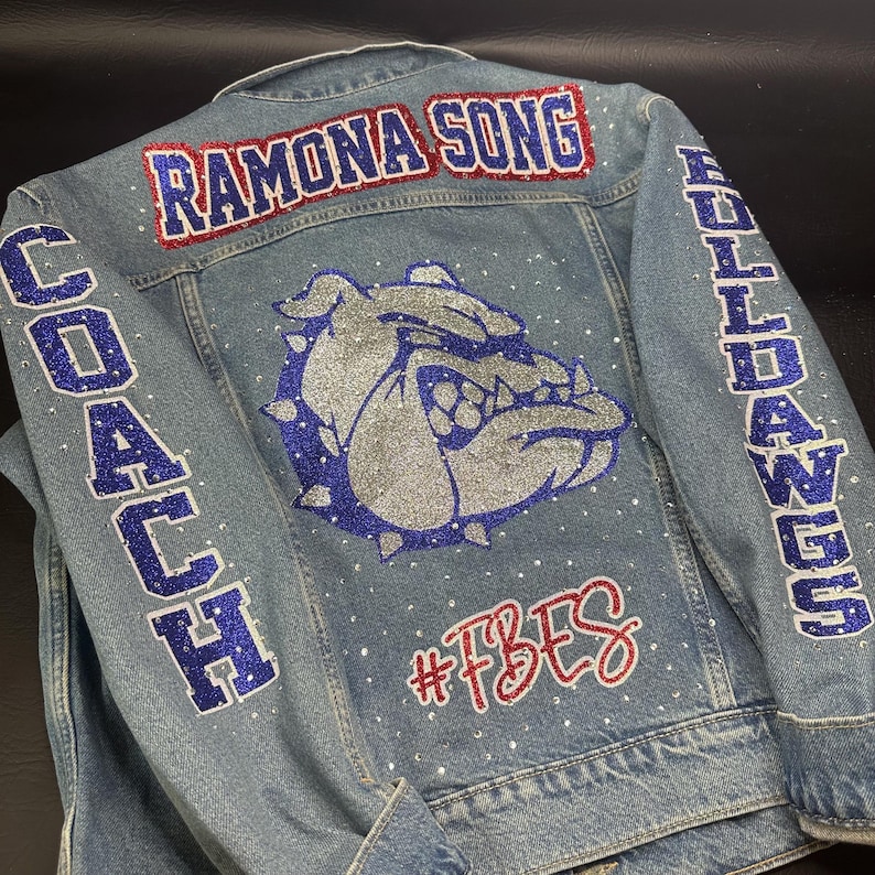 May include: A light blue denim jacket featuring a large silver and blue glitter bulldog graphic on the back. The text RAMONA SONG is in red and blue glitter, COACH and BULLDOGS are in blue glitter, and #FBES is in red glitter. Small clear rhinestones are scattered.