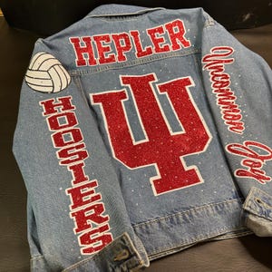 May include: Light blue denim jacket with red glitter lettering and accents. Features the name "HEPLER" across the shoulders, the word "HOOSIERS" down the left sleeve, and the Indiana University logo on the back. A volleyball patch is on the left shoulder.