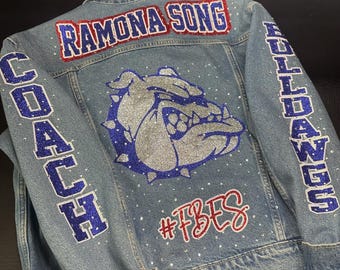 May include: A light blue denim jacket featuring a large glittery silver and blue bulldog graphic on the back. The text RAMONA SONG is in red and blue glitter, COACH and BULLDOGS are in blue glitter, and #FBES is in red glitter. Small clear rhinestones are scattered throughout.