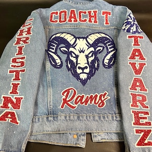 May include: Light blue denim jacket with a large ram head graphic in navy and white. The jacket features the text "COACH T" on the back, "Rams" below the ram, and the name "CHRISTINA" on one sleeve and "TAVAREZ" on the other sleeve in red glitter.