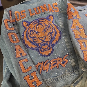 May include: A blue denim jacket featuring a large tiger head graphic in orange and purple glitter. The text LOS LUNAS, COACH, TIGERS, and AMANDA is applied in orange glitter lettering. The jacket is embellished with scattered clear rhinestones.