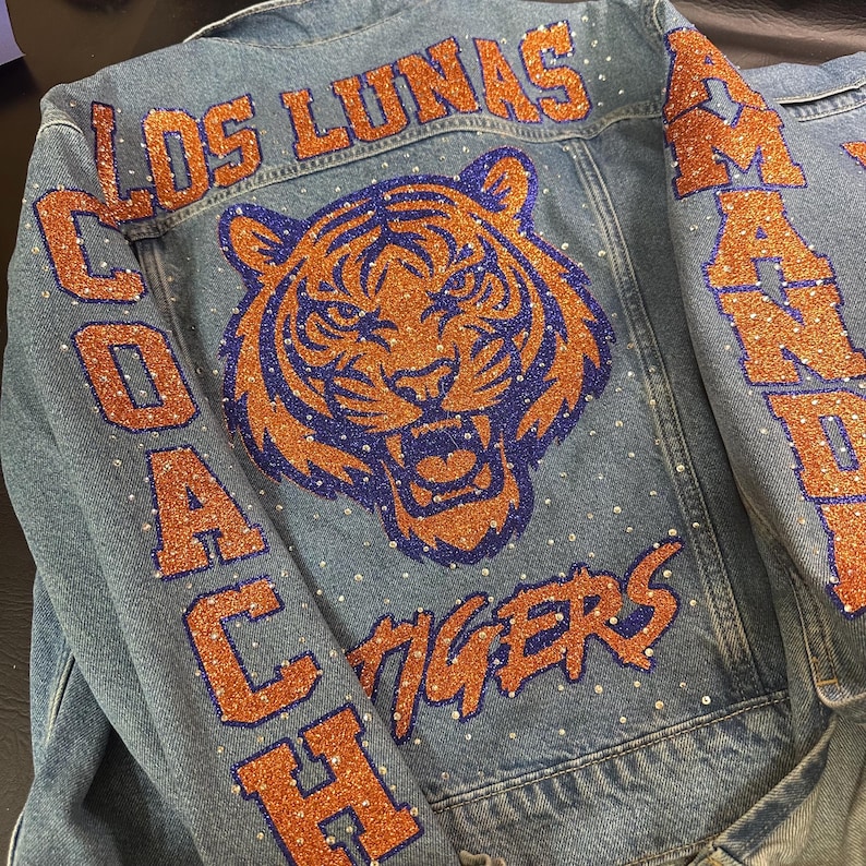 May include: A blue denim jacket featuring a large tiger head graphic in orange and purple glitter. The text LOS LUNAS, COACH, TIGERS, and AMANDA is applied in orange glitter lettering. The jacket is embellished with scattered clear rhinestones.