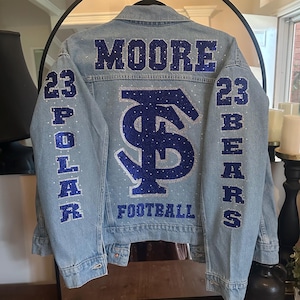 May include: Light blue denim jacket with the word "MOORE" in blue glitter across the back shoulders. The jacket has a large blue glitter "FS" logo and the word "FOOTBALL" below it. The sleeves have the numbers "23" and the words "POLAR" and "BEARS" in blue glitter.