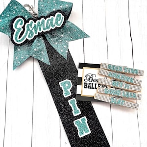 May include: A teal and black cheer bow with the name "Esmae" on it. The bow is attached to a black and teal cheer pin with the word "PIN" on it. There are also five teal and silver cheer clips with the word "PIN" on them.