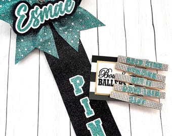 Peut inclure: Un nœud de pom-pom de cheerleading bleu sarcelle et noir avec le nom "Esmae" dessus. Le nœud est attaché à une épingle de cheerleading noire et bleu sarcelle avec le mot "PIN" dessus. Il y a aussi cinq pinces de cheerleading bleu sarcelle et argent avec le mot "PIN" dessus.